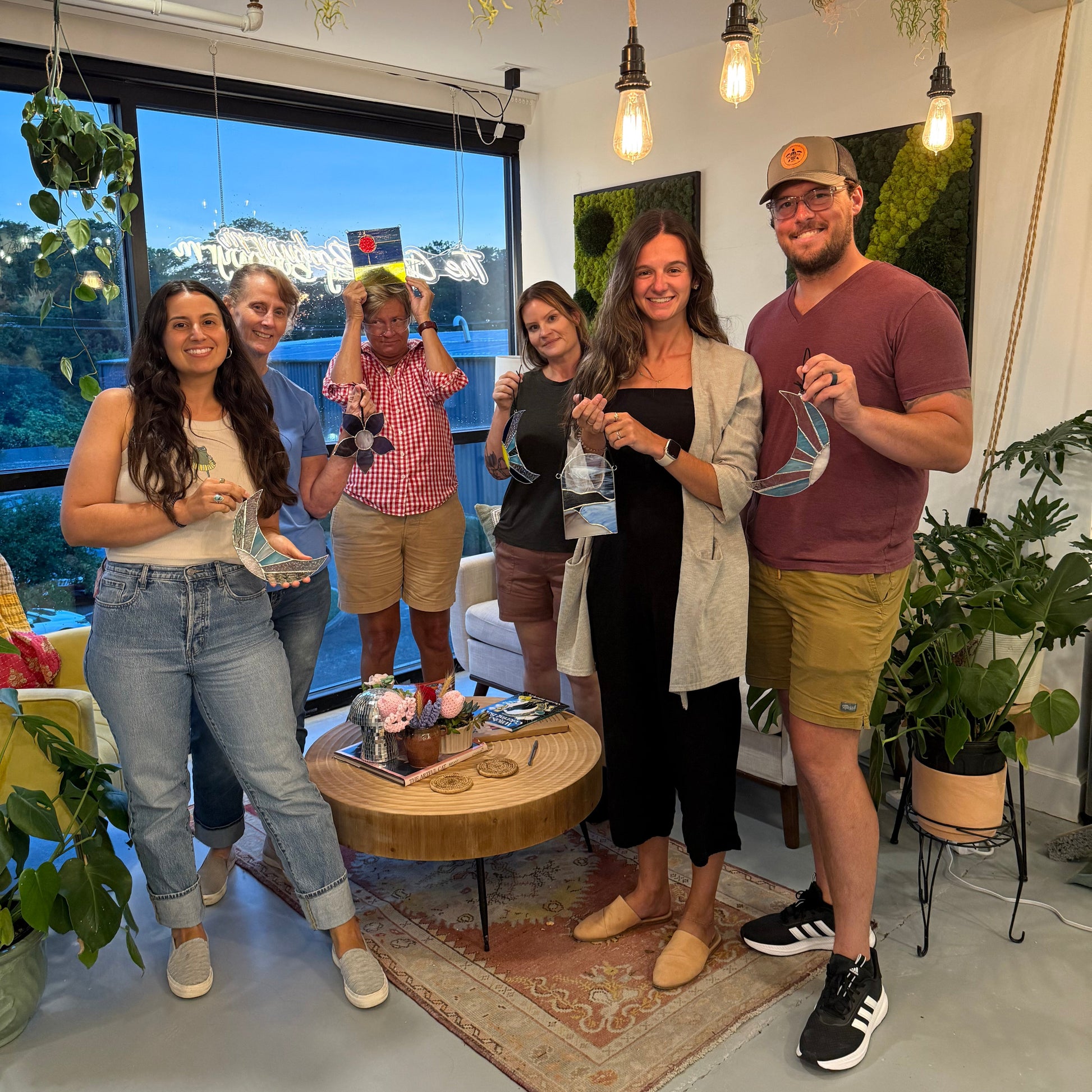 Group of people posing together after a stained glass workshop in a casual indoor setting with plants and a large window in the background.